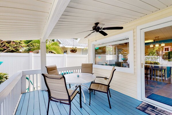 Une terrasse extérieure couverte, dotée d’un ventilateur de plafond, d’une table ronde avec des chaises, et offrant une vue sur la piscine grâce à une porte vitrée.