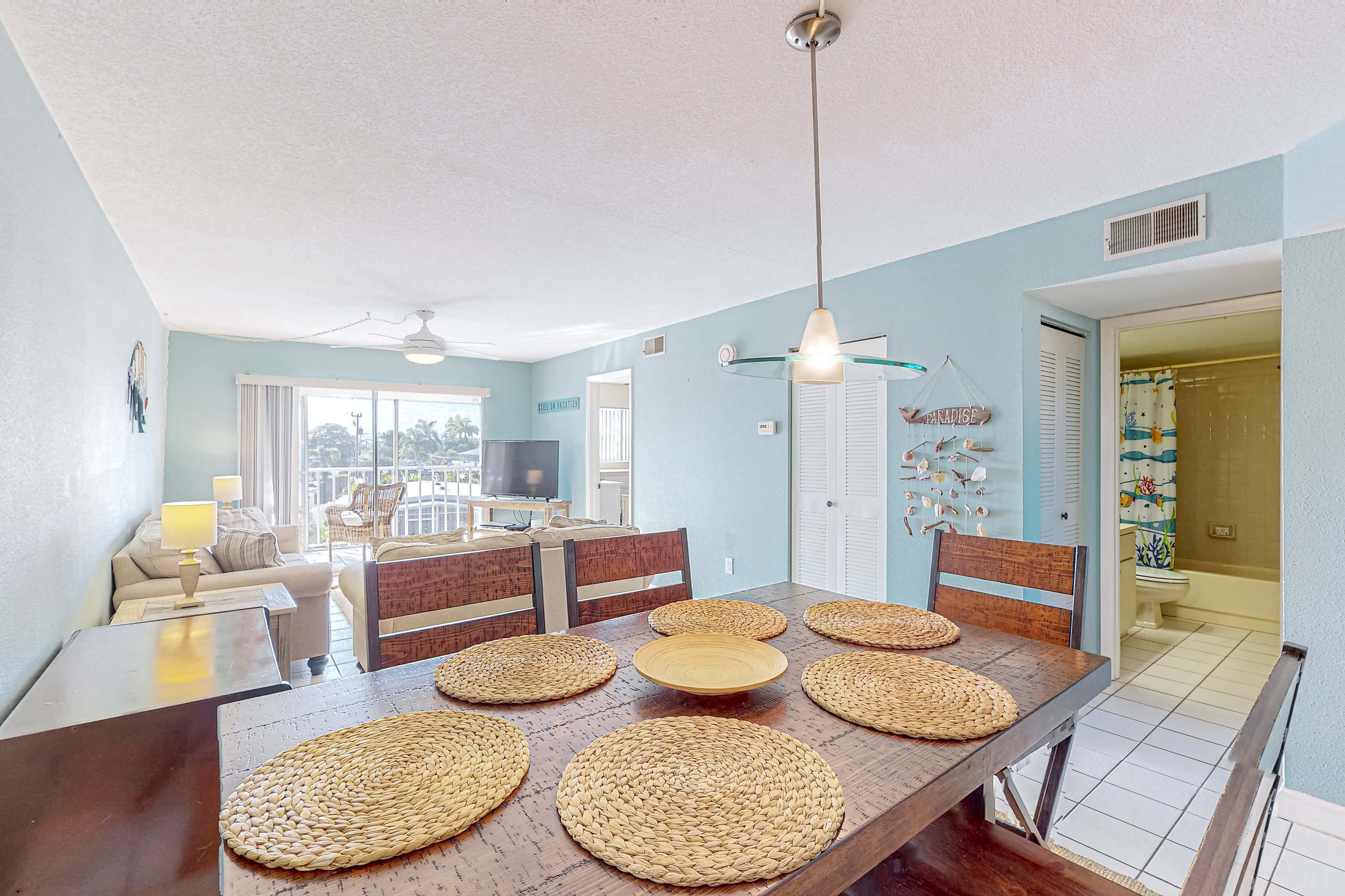 A dining area with a wooden table set with woven placemats, a ceiling fan, and a view of the living room and balcony.