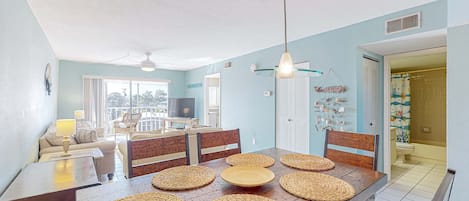 A dining area with a wooden table set with woven placemats, a ceiling fan, and a view of the living room and balcony.