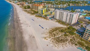 Vista aérea de un hotel frente a la playa con varias edificaciones, una playa de arena y una masa de agua.