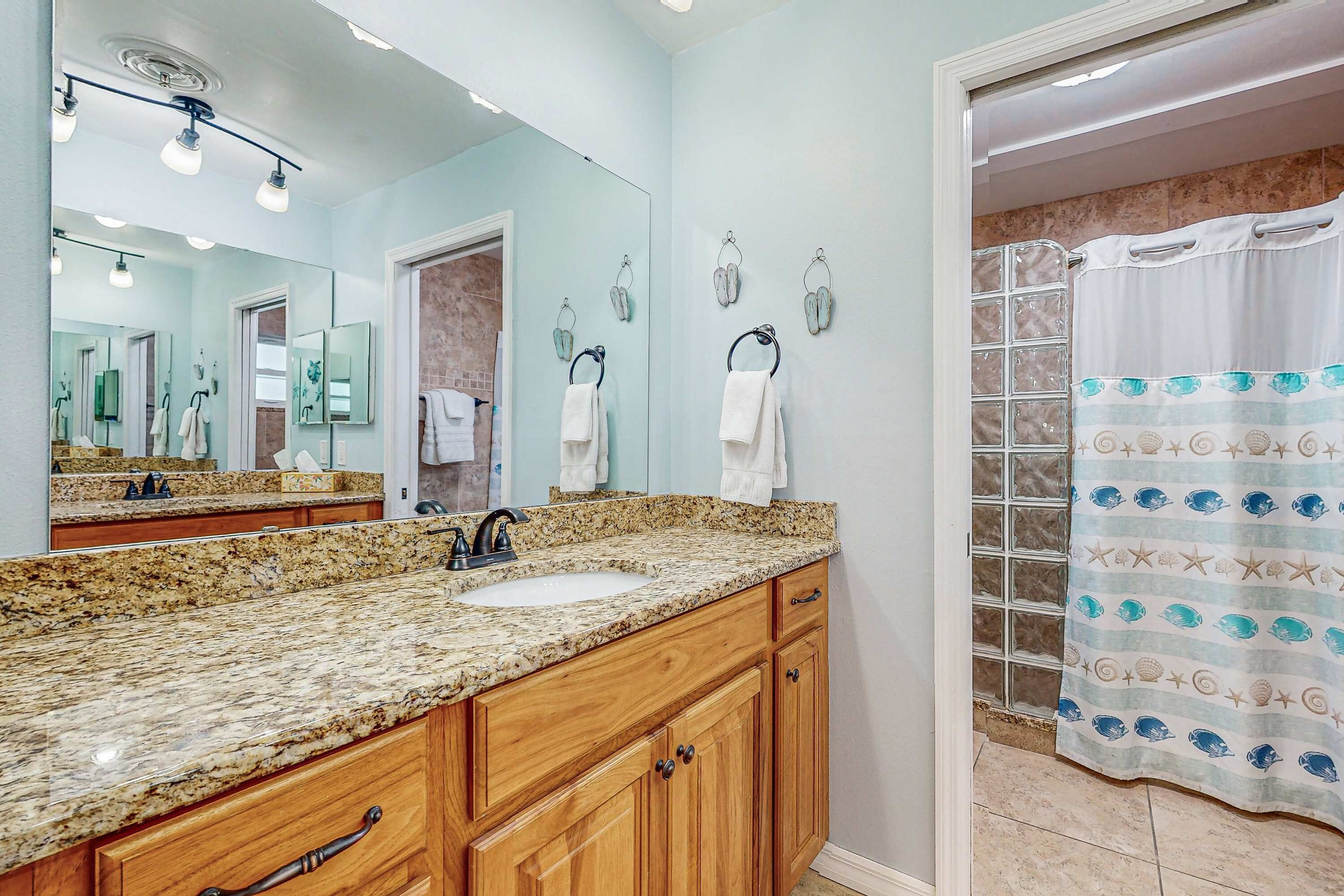 A bathroom with a granite countertop sink, wooden cabinets, a large mirror, and a shower with a patterned curtain.