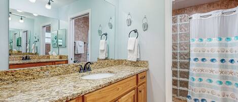 A bathroom with a granite countertop sink, wooden cabinets, a large mirror, and a shower with a patterned curtain.