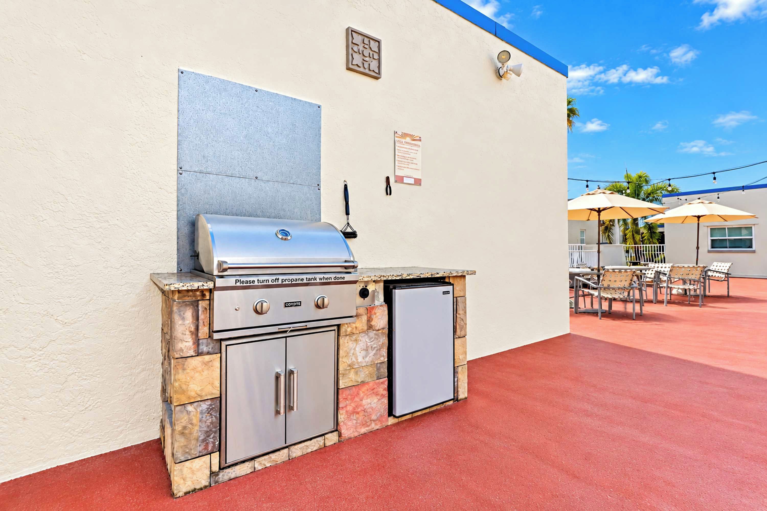 Un espacio al aire libre con una parrilla y refrigerador, un patio con sillas y sombrillas, y un cielo despejado.