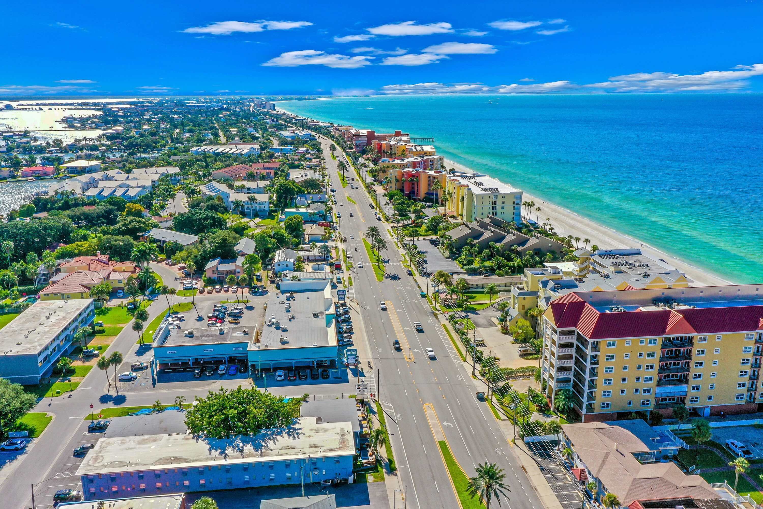 Vista aérea de uma cidade costeira com um hotel em destaque à beira-mar, ruas largas e águas azul-claras.