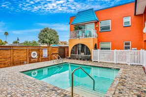 Ein Schwimmbecken mit Holzterrasse, umzäunter Bereich und Gebäude mit blauem Dach und orangen Wänden.