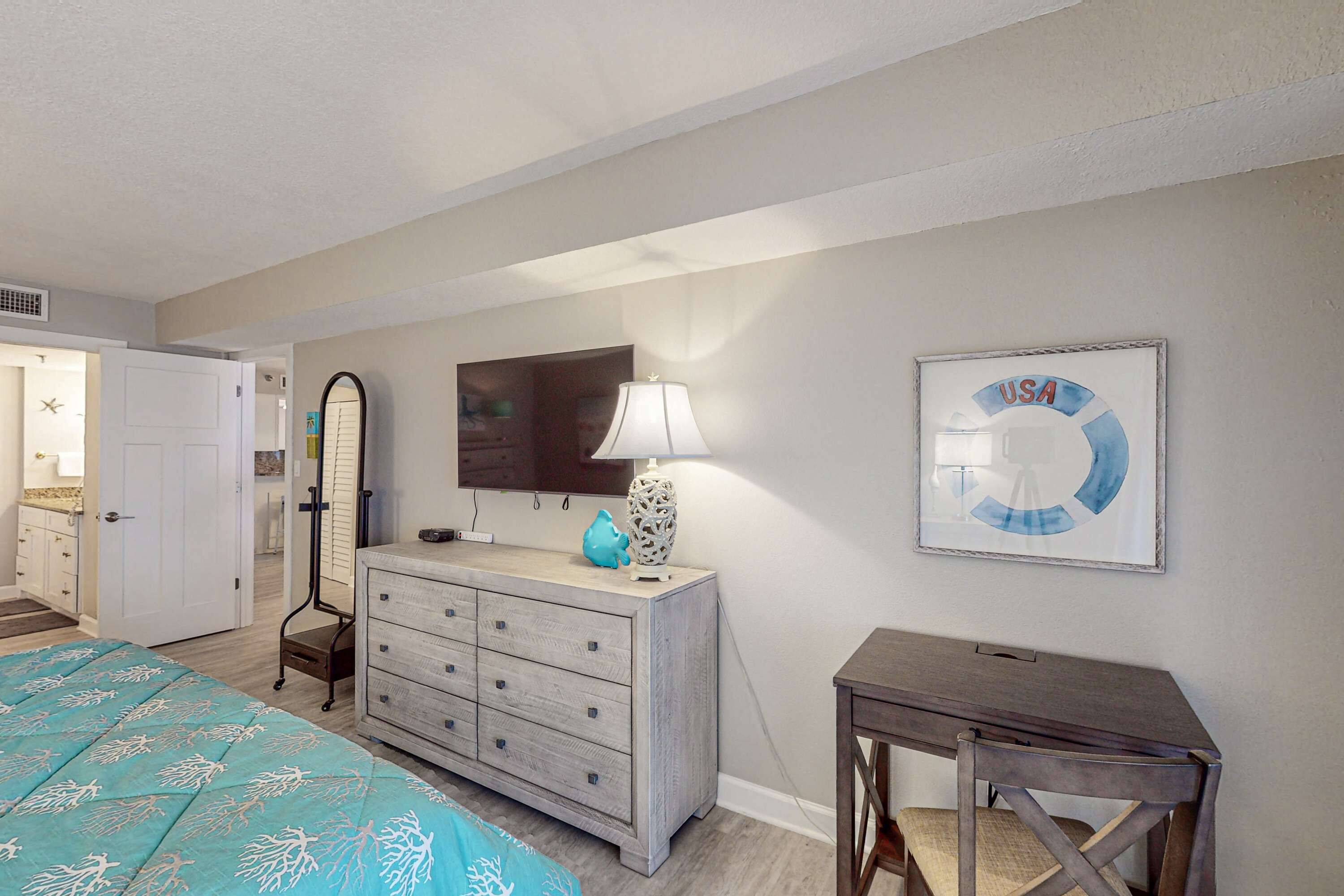 A bedroom with a bed, dresser, vanity, mirror, lamp, and a wall with a framed picture.