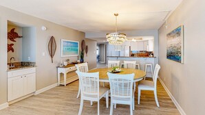 A dining area with a wooden table and white chairs, a chandelier, and a kitchen in the background. - Renovated beachfront condo with heated pool amazing Gulf views and washer dryer (Madeira Beach)