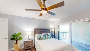Ein Schlafzimmer mit einem Bett, einem Deckenventilator, einem Nachttisch, einer Lampe, einem Blumentopf und Blick auf einen Balkon.