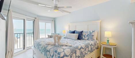A bedroom with a bed, a ceiling fan, a nightstand, and a view of the beach.