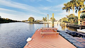 A wooden dock with a picnic table and chairs, a calm river, and a row of palm trees in the background.