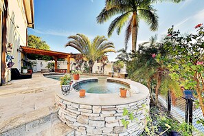 A stone pool surrounded by potted plants and palm trees, with a covered seating area and a pathway leading to a building.