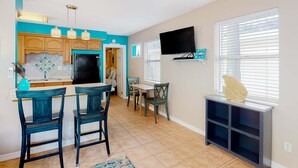 A kitchen with a black refrigerator, wooden cabinets, and a turquoise backsplash. There is a dining area with a table and chairs, and a TV mounted on the wall. - Charming single level cottage with a kitchen enclosed yard and WiFi (Treasure Island)