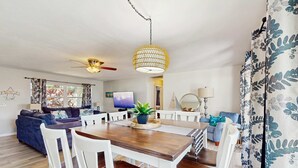 A modern living room with a wooden dining table, white chairs, a ceiling fan, and a large window with curtains. - Inviting ranch home near the beach Florida room alfresco dining (Clearwater)