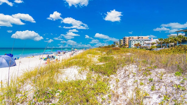 Uma praia com areia branca, céu azul límpido e alguns edifícios ao fundo.