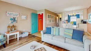 Una sala de estar con sofá, una mesa de centro y una mesita auxiliar con faroles. Se observa una puerta con marco colorido, un área de cocina y una mesa de comedor con sillas.