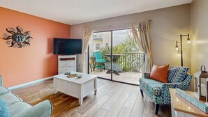 Una sala de estar con una televisión de pantalla plana, una mesa de centro blanca, un sofá estampado y un balcón con mesa y sillas.