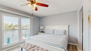Une chambre avec une grande fenêtre, un ventilateur de plafond, un lit avec une tête de lit blanche et un banc.