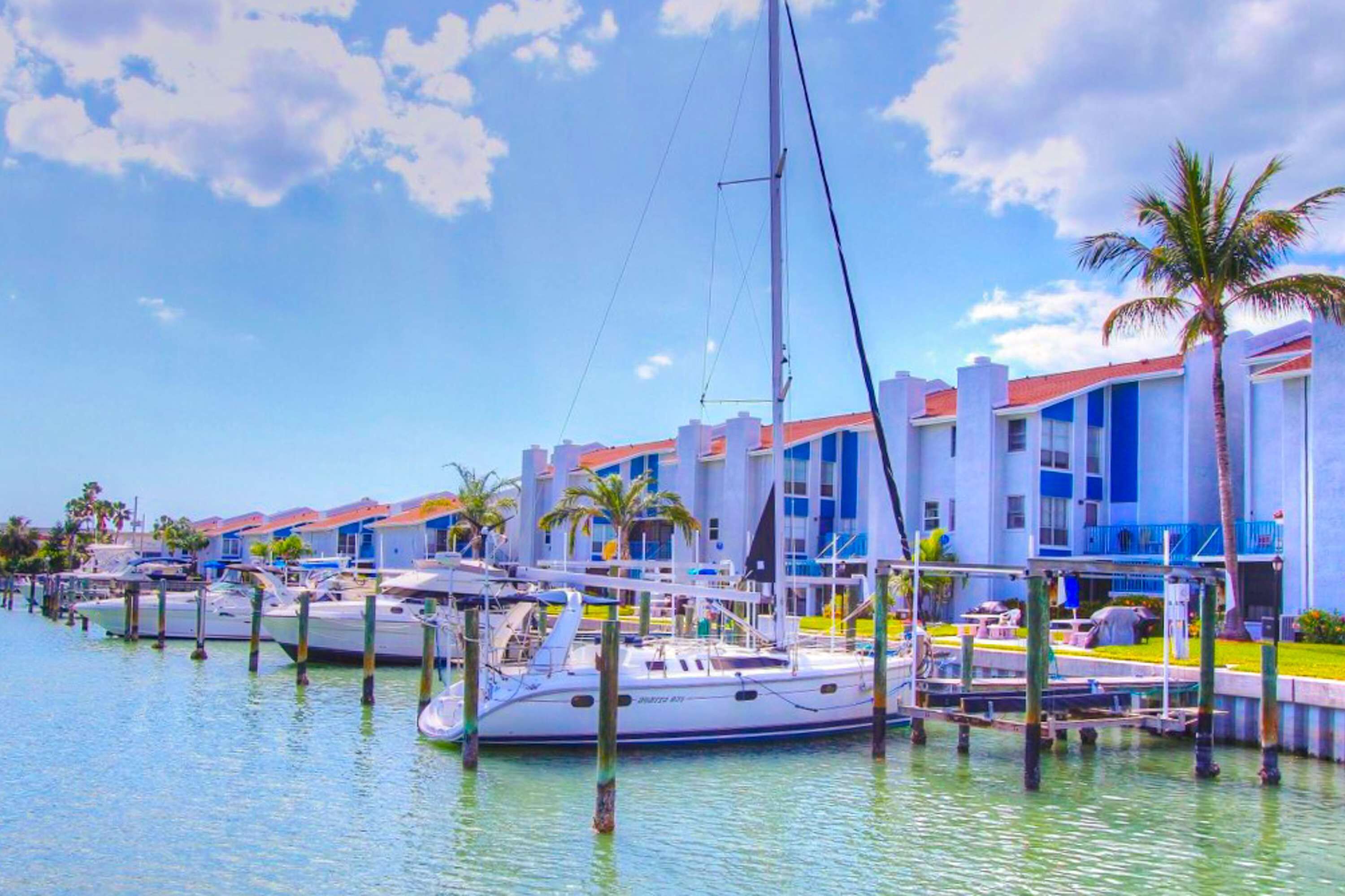 Un puerto deportivo con barcos amarrados, un edificio de fachada azul y blanca y palmeras.