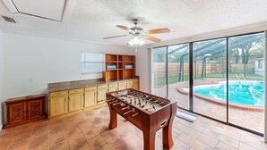 A spacious room with a foosball table, a ceiling fan, and a large sliding glass door leading to a backyard with a swimming pool.