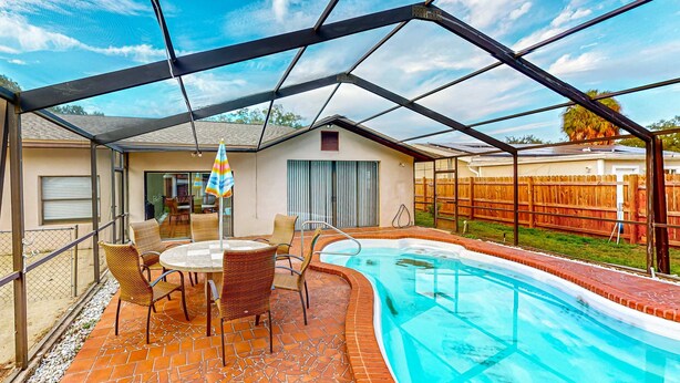 A covered outdoor swimming pool area with a patio table and chairs, a fenced yard, and a clear sky.
