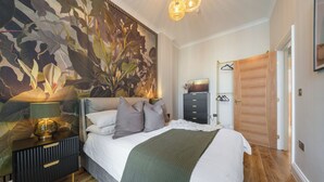 A bedroom with a large floral mural, a bed with a white comforter and green throw, a nightstand with a lamp, and a wooden door. - Cambridge Garden Apartments by Argyl (London)