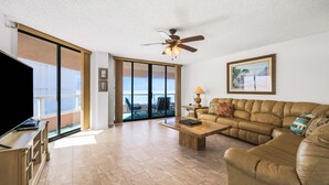 A living room with a sofa, coffee table, TV, and a ceiling fan. - Beachfront condo with multiple balconies heated pool gym hot tub and AC (Clearwater Beach)