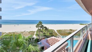 A balcony with a view of a beach, palm trees, and a pool area. - Beachfront condo with multiple balconies heated pool gym hot tub and AC (Clearwater Beach)