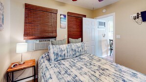 A bedroom with a bed, a nightstand with a lamp, a ceiling fan, and a door leading to another room.