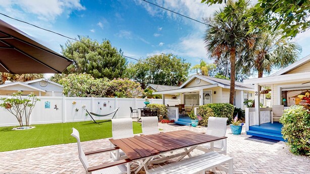 A patio area with a wooden table and white chairs, surrounded by a white fence and greenery.
