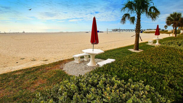 A beachside area with white tables and red umbrellas, palm trees, and a clear sky.