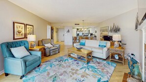 A living room with a sofa, armchair, coffee table, and a patterned rug. There are framed pictures on the wall, a chandelier, and a kitchen in the background.