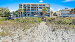 Un hotel frente a la playa con varios balcones, un paseo marítimo de arena y abundante vegetación.