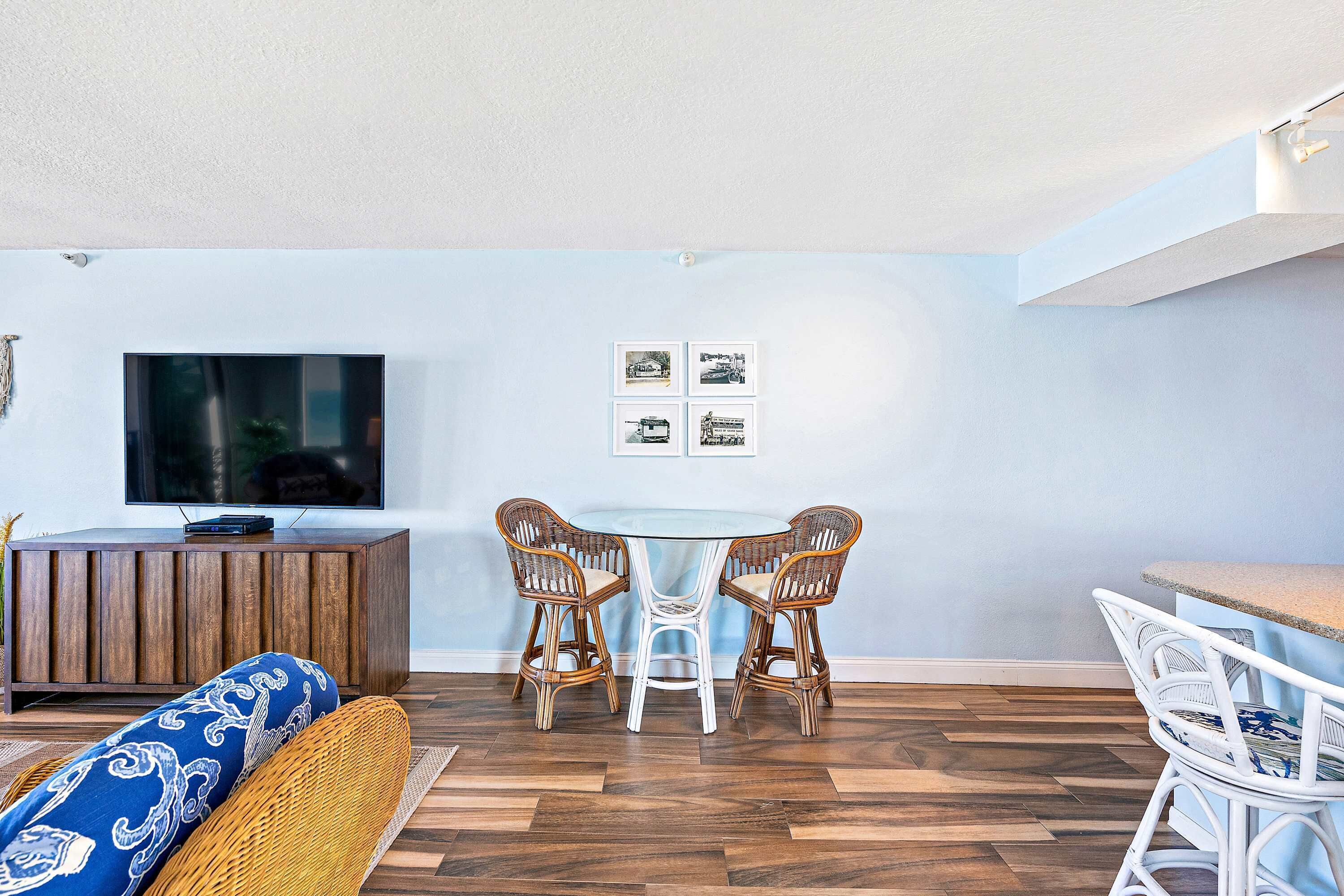 A modern living room with a round dining table, wicker chairs, a sofa with patterned cushions, a flat-screen TV, and a wooden cabinet.