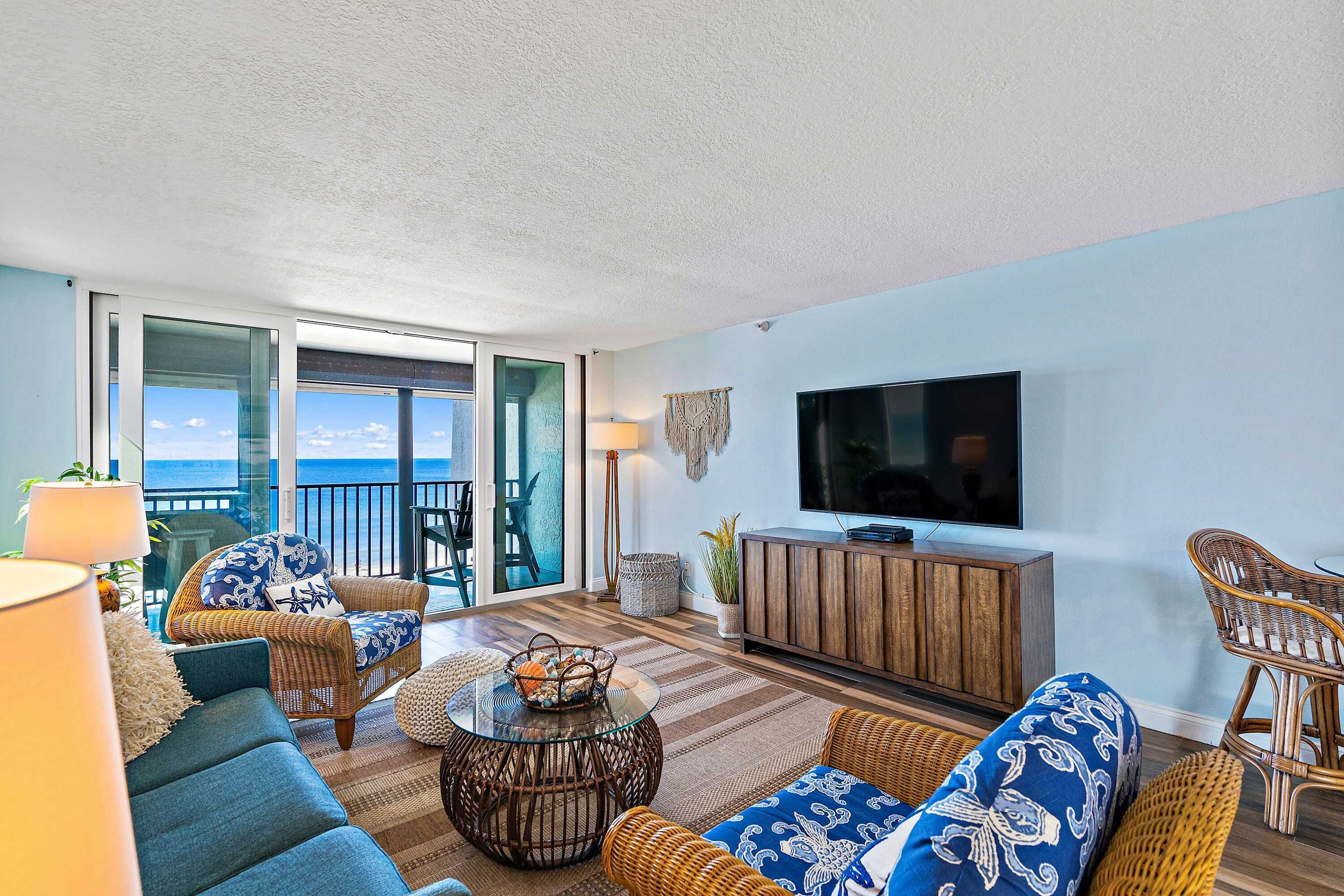 A living room with a large TV, wicker furniture, and a view of the ocean.