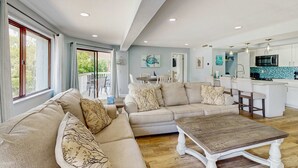 A spacious living room with a beige sofa set, a wooden coffee table, and a kitchen area with white cabinets and a blue backsplash. - Gulf side condo with heated pool hot tub central AC and WD snowbird friendly (Indian Shores)