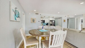 A dining area with a wooden table and white chairs, a kitchen in the background, and a living area with a sofa. - Gulf side condo with heated pool hot tub central AC and WD snowbird friendly (Indian Shores)
