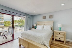 A bedroom with a bed, a nightstand, a lamp, a window with a view of buildings and greenery, and a balcony with chairs.