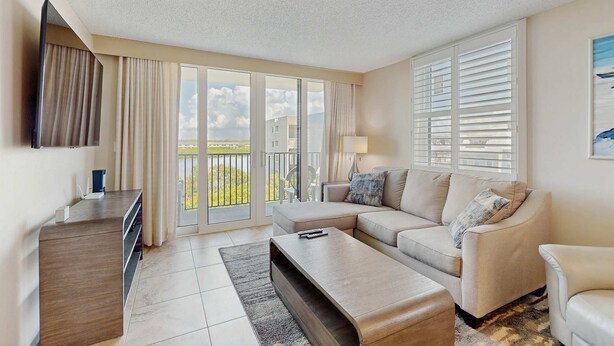 A modern living room with a sofa, a coffee table, and a TV on a wooden stand. There is a balcony with curtains and a view of the outside.