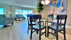 A modern living room with a dining area, featuring a ceiling fan, large windows, and a painting on the wall. - 4th floor beachfront condo with private balcony views pool BBQ and AC (Indian Shores)