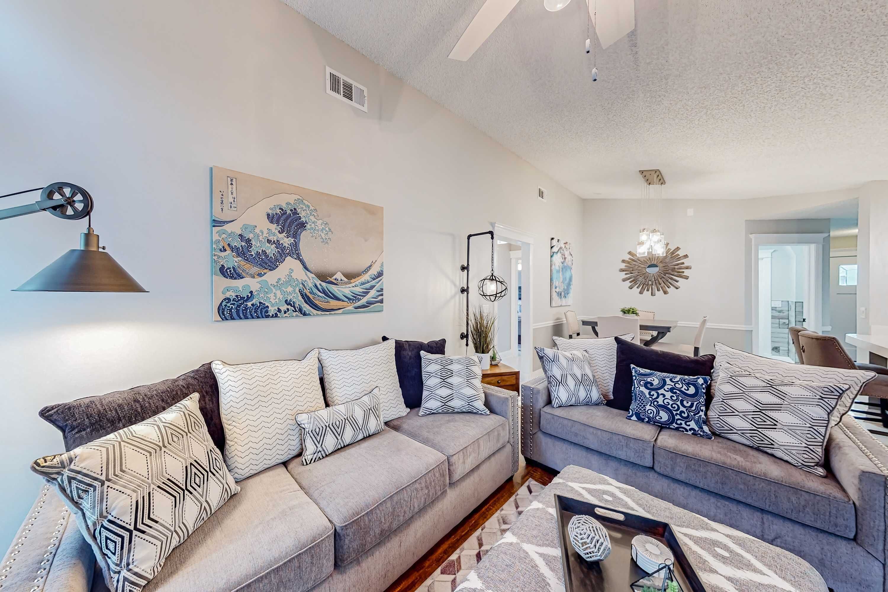 A living room with two grey sofas, patterned cushions, a wall-mounted lamp, and a large wave painting.