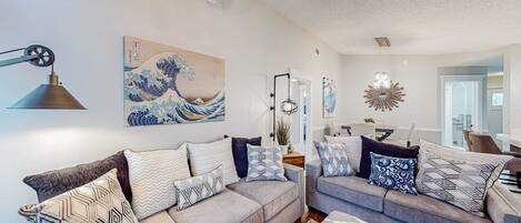 A living room with two grey sofas, patterned cushions, a wall-mounted lamp, and a large wave painting.