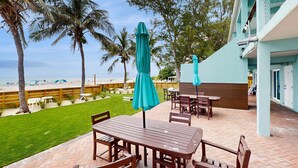 Une terrasse extérieure avec des tables et des chaises en bois, une pelouse verdoyante et une vue sur la plage et les palmiers.