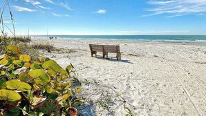 Ein Sandstrand mit einer Holzbank, blauem Himmel und Personen in der Ferne.