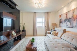 A modern living room with a flat-screen TV, a sofa, a coffee table, and a large abstract painting. - Apto c/ piscina e churrasq, 280m da praia IMD0502 (Bombinhas)