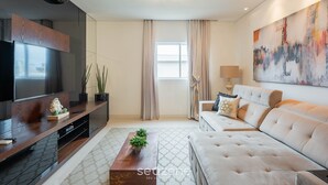 A modern living room with a flat-screen TV, a sofa, a coffee table, and a large abstract painting. - Apto c/ piscina e churrasq, 280m da praia IMD0502 (Bombinhas)