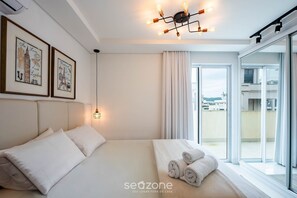 A modern bedroom with a large bed, a ceiling light fixture, and a view of the city through the window. - Apto c/ piscina e churrasq, 280m da praia IMD0502 (Bombinhas)