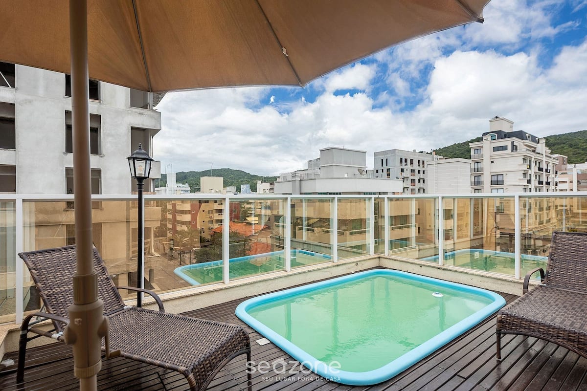 Ein Poolbereich auf dem Dach mit Korbstühlen und einem großen Sonnenschirm, umgeben von Glasgeländern und Gebäuden.