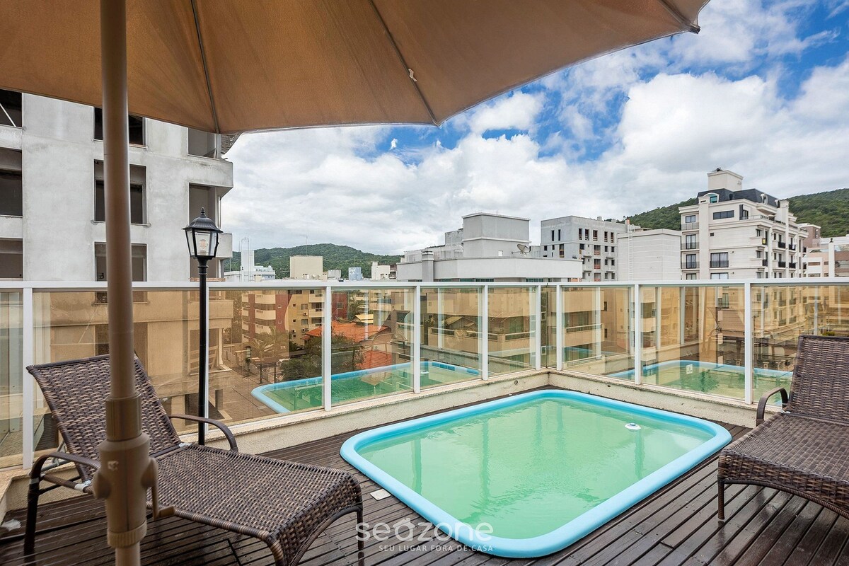 A rooftop pool area with wicker chairs and a large umbrella, surrounded by glass railings and buildings.