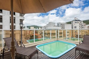 A rooftop pool area with wicker chairs and a large umbrella, surrounded by glass railings and buildings. - Apto c/ piscina e churrasq, 280m da praia IMD0502 (Bombinhas)