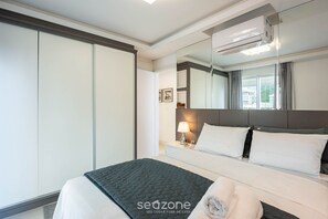 A hotel room with a bed, a nightstand, a lamp, a large mirror, and an air conditioning unit. - Apto c/ piscina e churrasq, 280m da praia IMD0502 (Bombinhas)